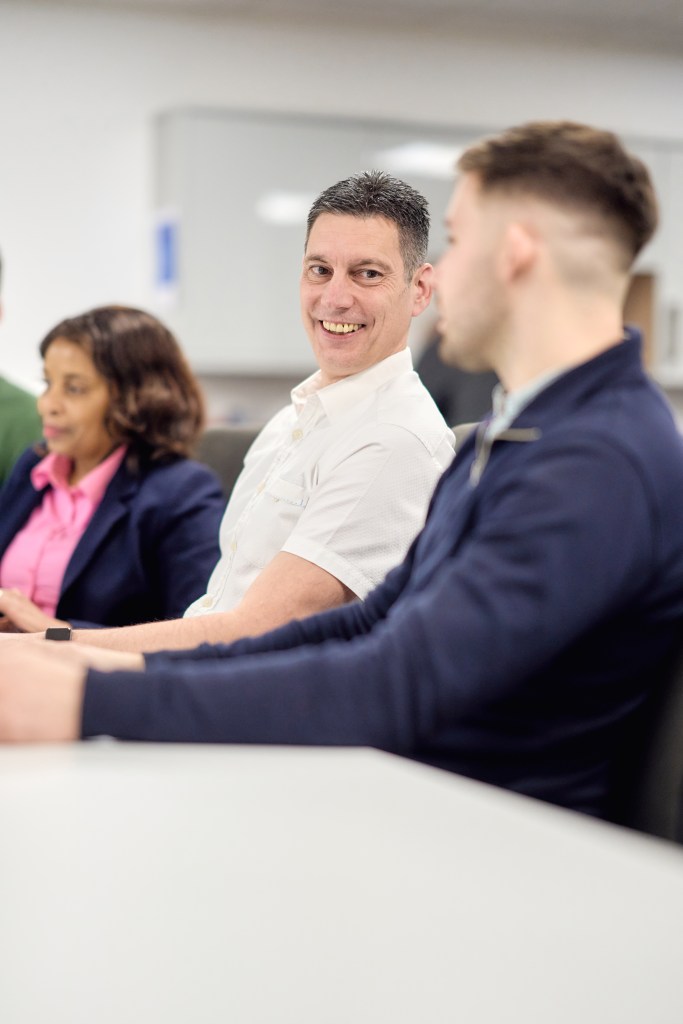 Three colleagues sitting at a table in a meeting, with one person smiling while talking to a coworker in a modern office.