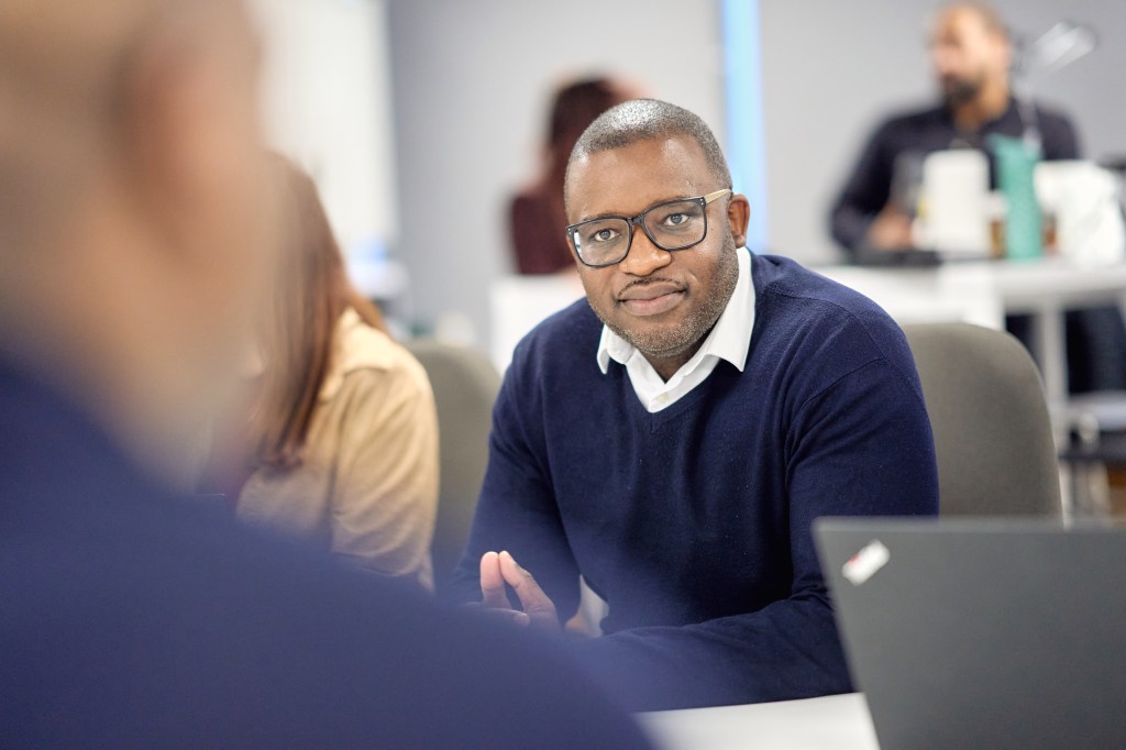 A colleague listening attentively during a meeting in a modern office, with laptops and other team members in the background.