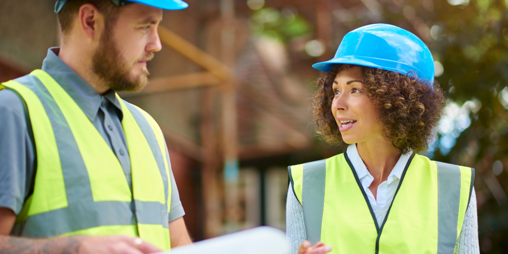 Two construction professionals wearing hard hats and high-visibility vests discussing plans at a building site.