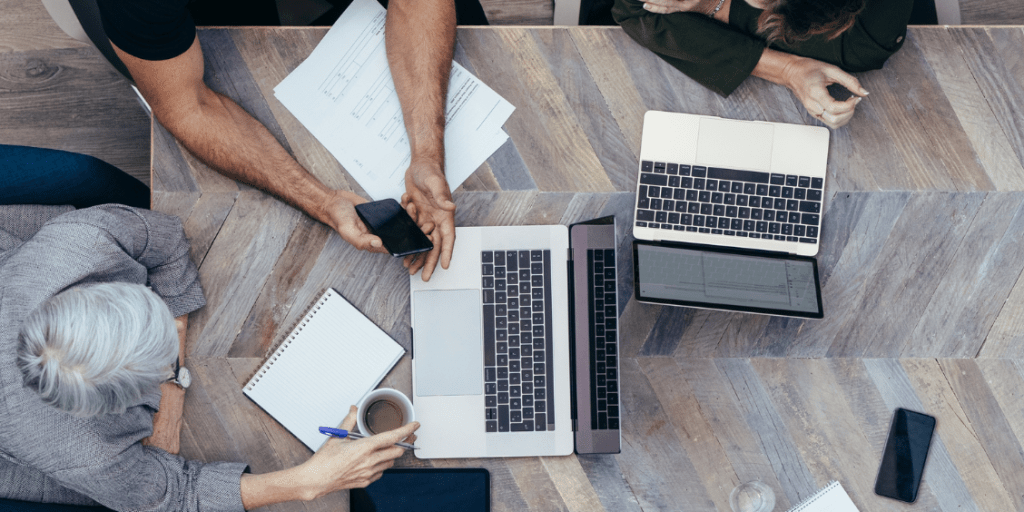 Overhead view of colleagues working together at a table with laptops, documents, notebooks and a smartphone during a meeting.