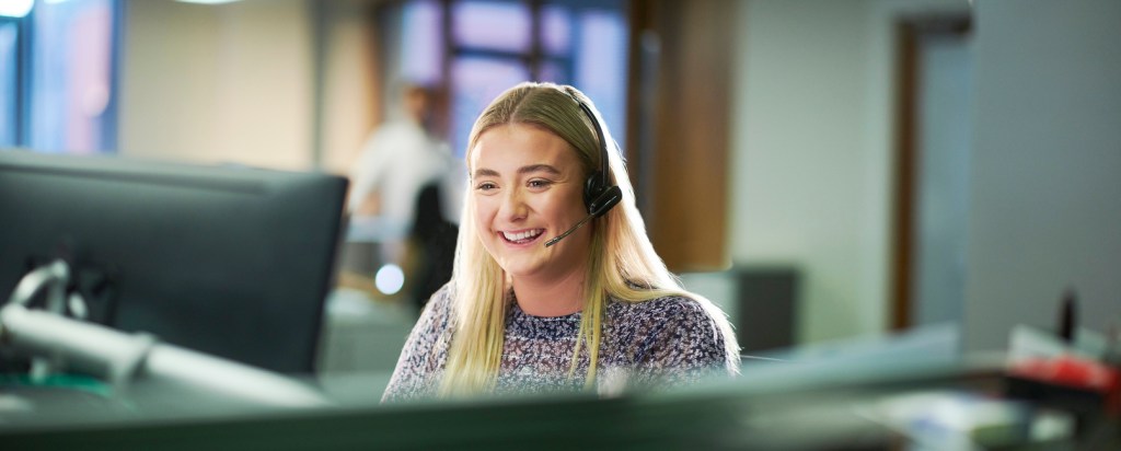 Smiling customer service representative wearing a headset, seated at a computer in a modern office environment.
