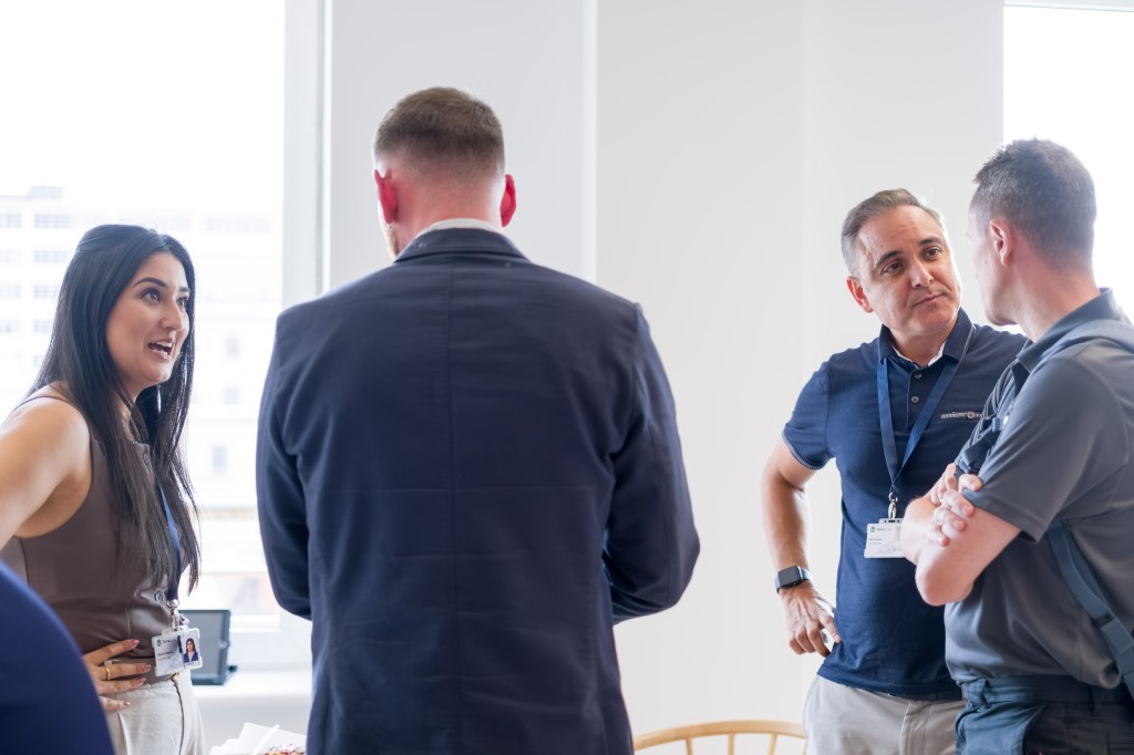 Four colleagues standing and talking in a bright office space, wearing lanyards and having a collaborative discussion near a window.