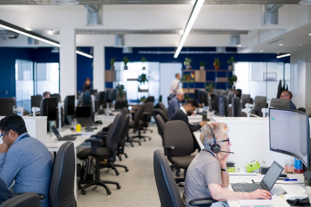 Colleagues working at computer desks in a modern open-plan office, with one person wearing a headset and using a laptop in the foreground.