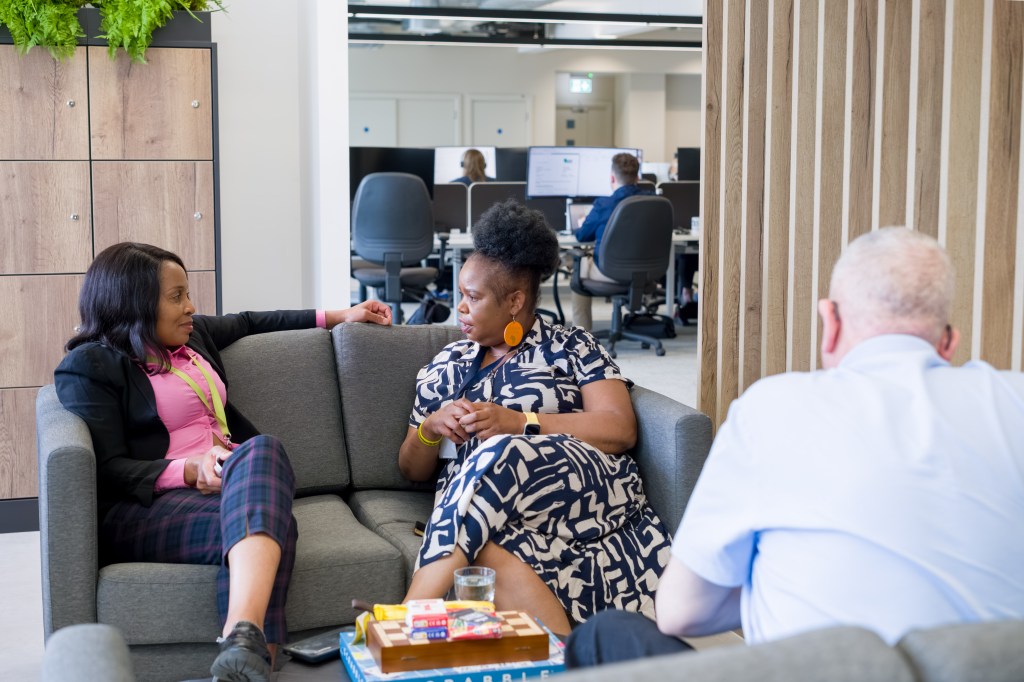 Three colleagues having an informal discussion on a sofa in a modern office breakout area, with desks and computer workstations visible in the background.