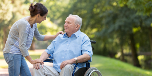 Lady leaning down talking to older man sat in wheelchair against garden backdrop