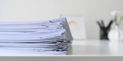 A stack of paper documents against a blurred white background with pots of pens