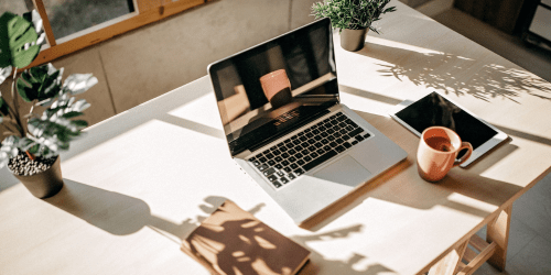 Laptop on modern desk in brightly lit room with coffee, notepad, tablet and plants