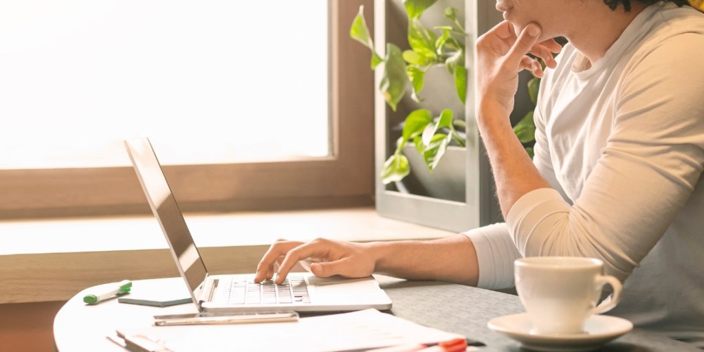 A man sits at a desk browsing on his open laptop by a bright window with a plant besides him and a coffee cup on the table beside him