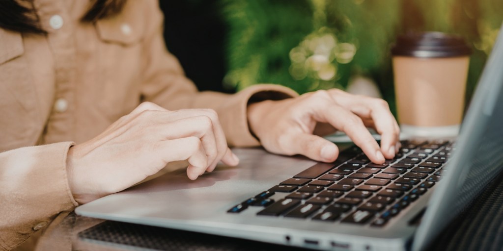 Shot of laptop user's hand as they type wearing a brown shirt with a disposable coffee cup on the desk next to them and blurred plants in the background