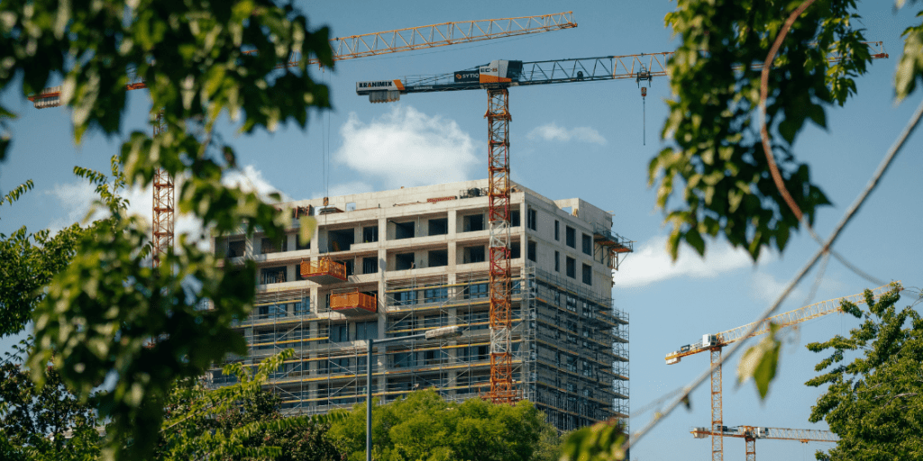Tower cranes constructing a high-rise building beneath a cloudy blue sky, with trees in the foreground.