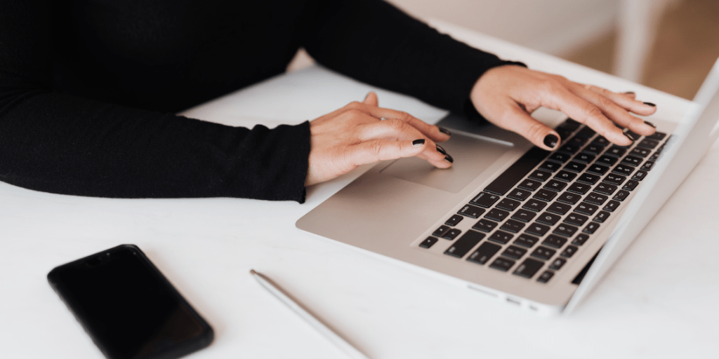 Hands typing on a laptop at a desk, with a smartphone and office workspace visibl