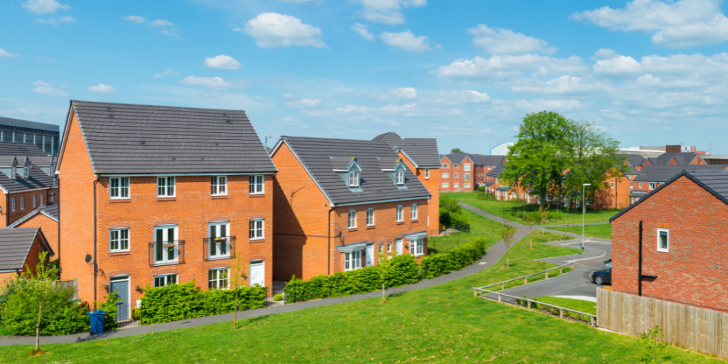 New-build red-brick houses on a residential estate, surrounded by green grass and trees beneath a bright blue sky with scattered clouds.