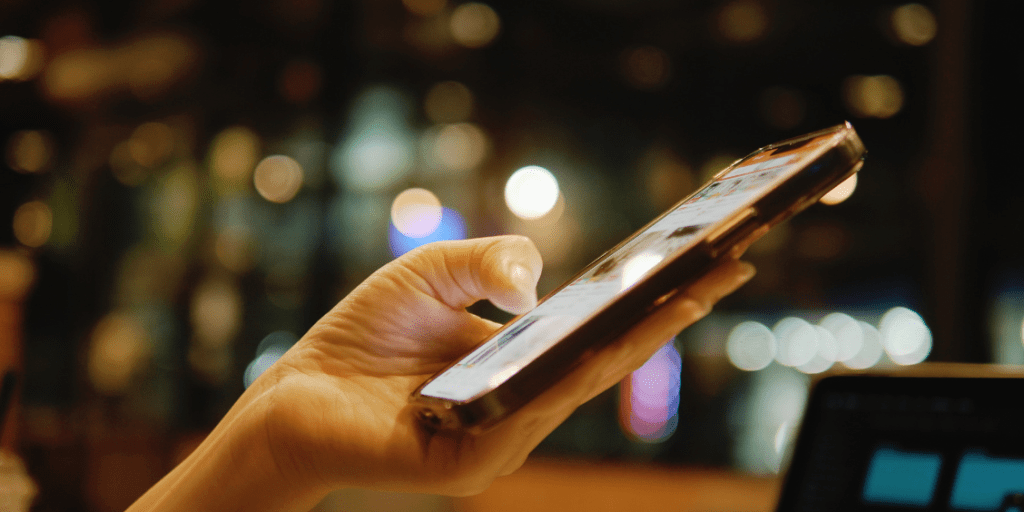 A hand scrolling on a smartphone against a softly blurred background of lights.