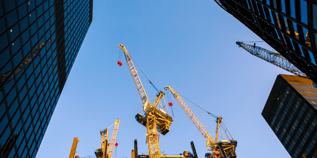 View of construction cranes between tall buildings against a clear blue sky.