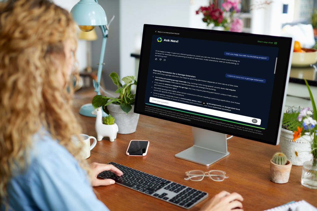 A person with curly hair is sitting at a desk using a computer. The screen displays a chat interface titled 'Ask Navi' with questions about building projects and planning permissions. The workspace is decorated with plants, a coffee mug, and a smartphone.