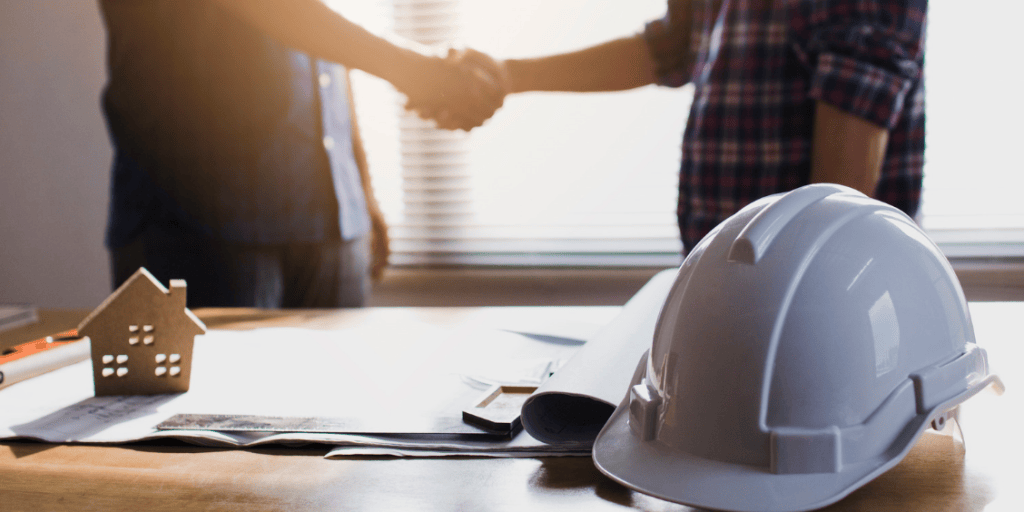 Architectural plans and a house model on a desk, with a hard hat in the foreground and two professionals shaking hands in the background.